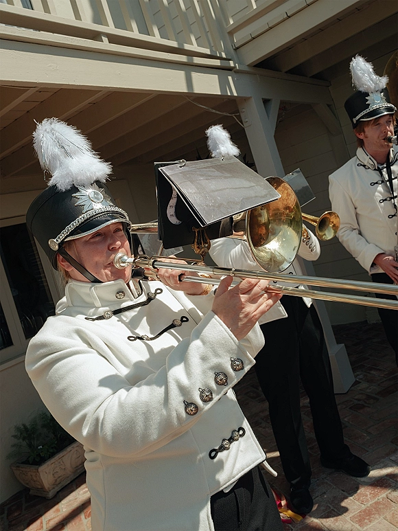 Wedding band in white uniforms playing brass instruments on a brick patio, trombone and trumpet visible with feather plumed hats in daylight