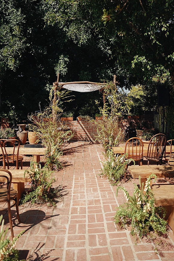 Ceremony setup for an outdoor wedding ceremony with a wooden arbor and fabric drape, greenery-lined brick aisle, benches on a garden patio