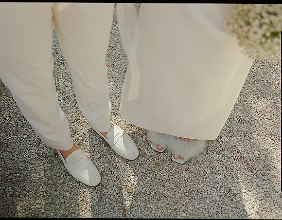Wedding shoe details with bride and groom shoes on sunlit gravel, white loafers beside heeled sandals and a feather hem dress in shadow