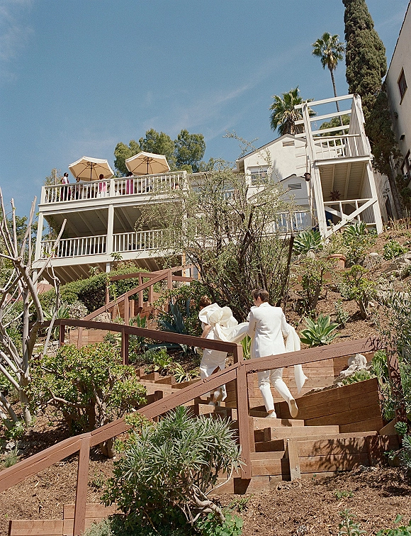 Wedding couple portrait of newlyweds walking upstairs hand in hand, bride’s gown train and groom suit on wooden steps in hillside garden backdrop