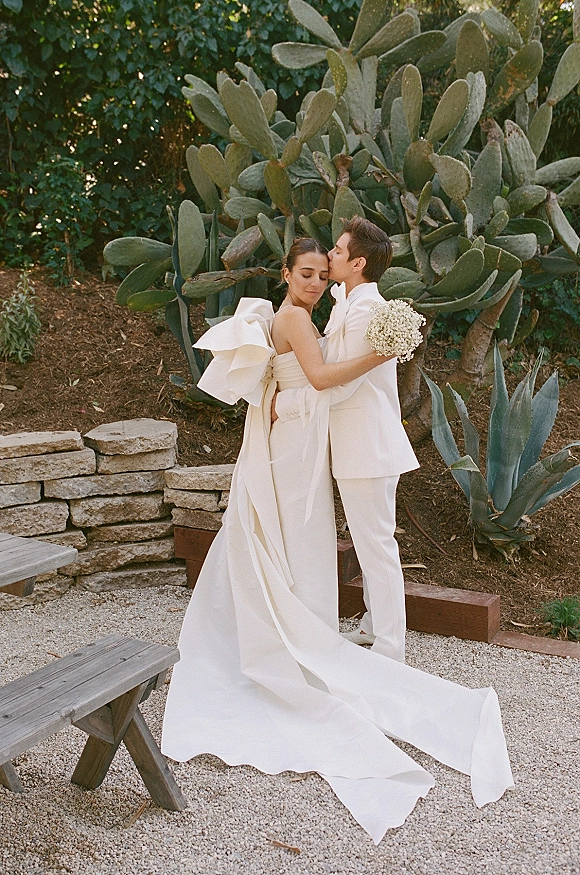 Couple portrait of bride and groom embrace with a forehead kiss, her shoulder-bow dress and long train in a cactus garden.