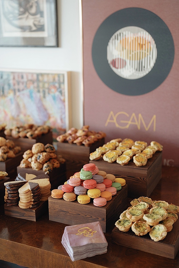 Wedding dessert table with a macaron dessert display of cookies and mini tartlets on wooden risers, with a printed sign against art prints wall