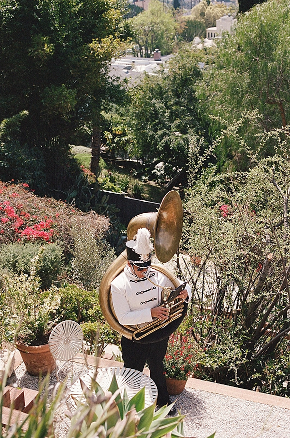 Wedding band musician with sousaphone wedding band uniform, reading sheet music on a stone patio amid garden greenery and hills backdrop