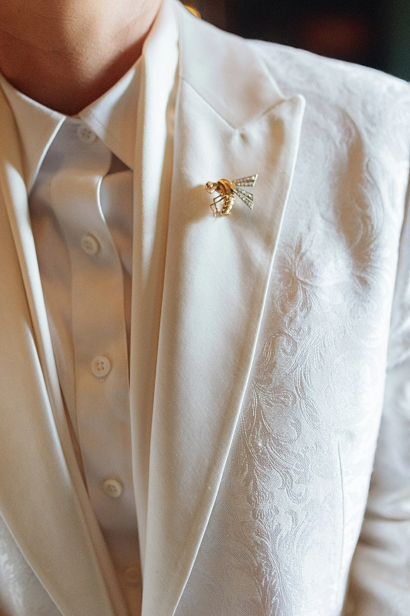 Groom suit details with a bee lapel pin on a white tuxedo jacket, waistcoat, and dress shirt in warm indoor light