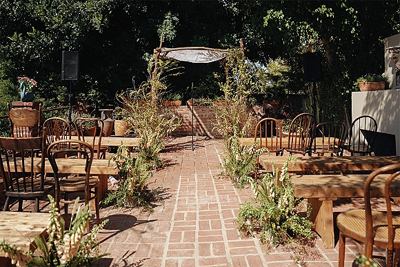 Ceremony setup with outdoor ceremony seating facing a rustic wooden altar arch, greenery aisle florals lining a brick patio courtyard