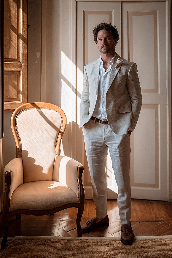 Groom portrait in a light suit with open-collar shirt and boutonniere, hands in pockets by paneled doors in window light