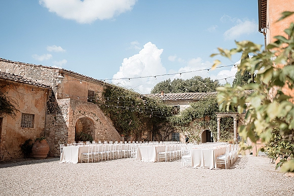 Reception tablescape with outdoor banquet tables in a U-shape, white linens and chairs, glassware, urn, and string lights in a stone villa courtyard