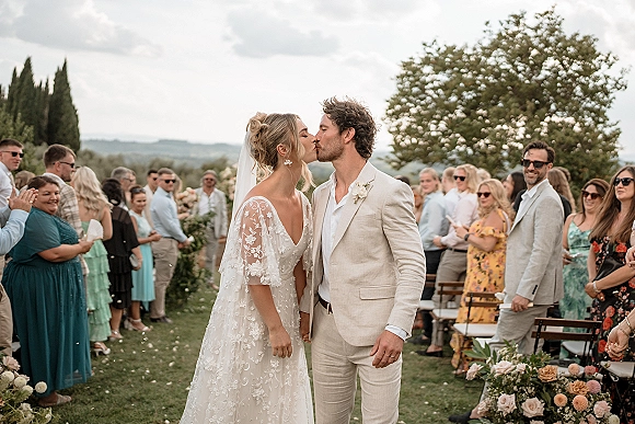 Wedding kiss as the couple shares a ceremony recessional kiss under a bride’s veil, guests cheering along a petal-strewn aisle on a lawn