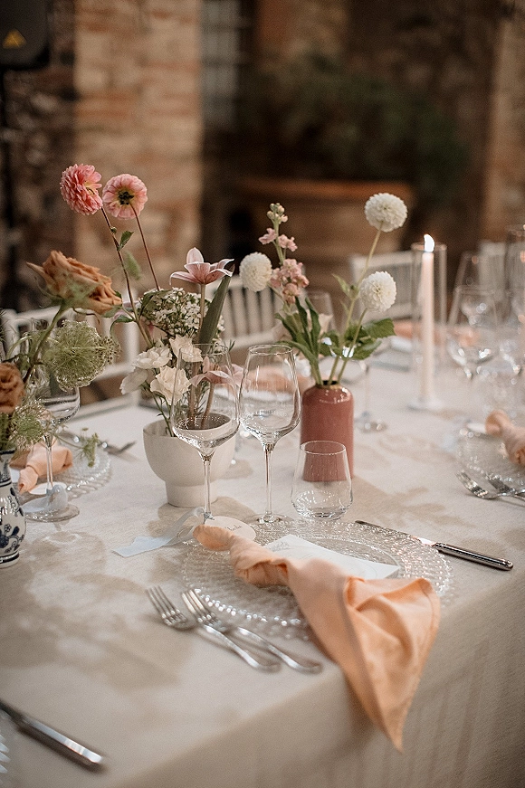 Reception tablescape with wedding table setting on white linen, bud vases of mixed florals and taper candles, brick wall behind