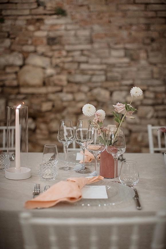 Reception tablescape wedding table setting with blush flowers in bud vases, taper candles in glass hurricanes, and place cards by a stone wall
