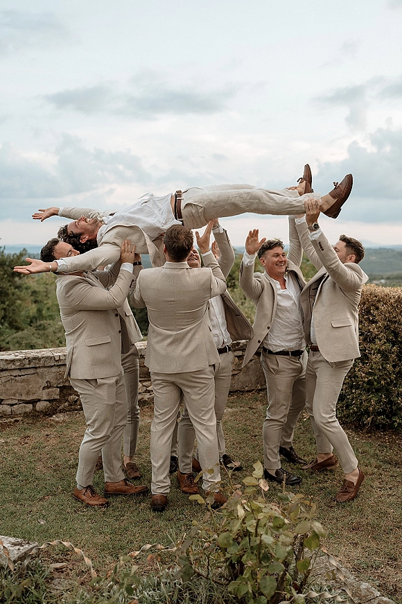 Groomsmen group photo as friends lift the groom, all in beige suits, laughing by a stone wall with cloudy hills behind