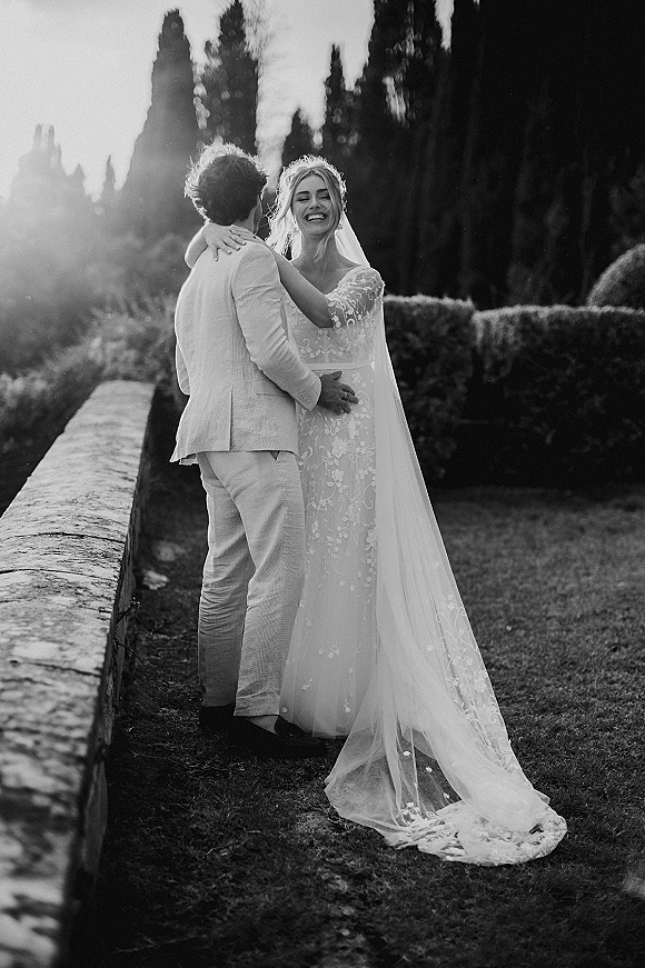 Couple portrait of bride and groom embracing, bride in lace gown and veil with groom in suit, laughing in a sunlit garden by a stone wall