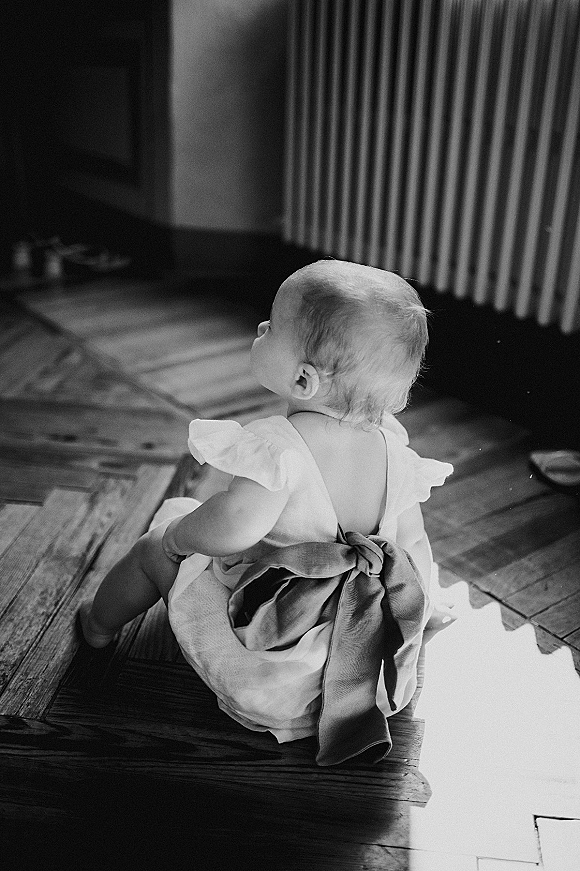 Flower girl in a flower girl dress sits on a wood floor by a radiator, soft window light highlighting her back bow sash
