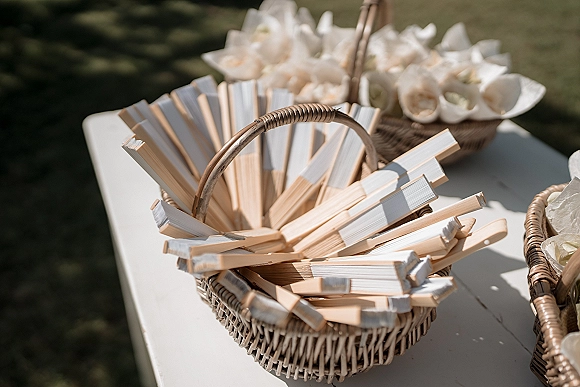 Wedding fan basket with wooden folding fans and white flower petals on a white table, sunlit on a grassy lawn with shadows