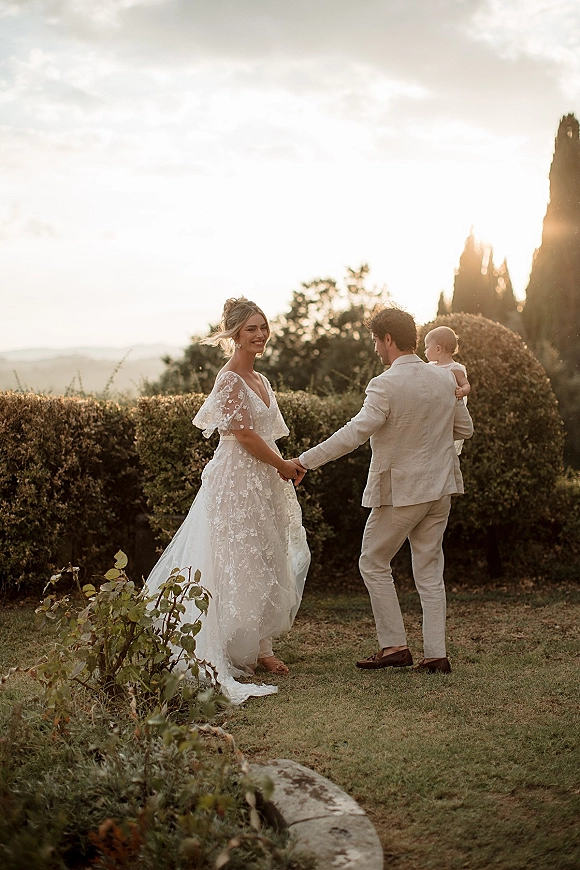 Couple portrait of bride and groom holding hands, walking away in a garden at sunset, her lace wedding dress flowing behind