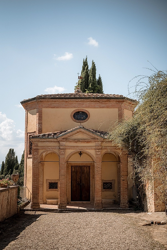 Chapel exterior with arched portico framing wooden double doors, terracotta roof tiles, and stone steps along a gravel path under blue sky