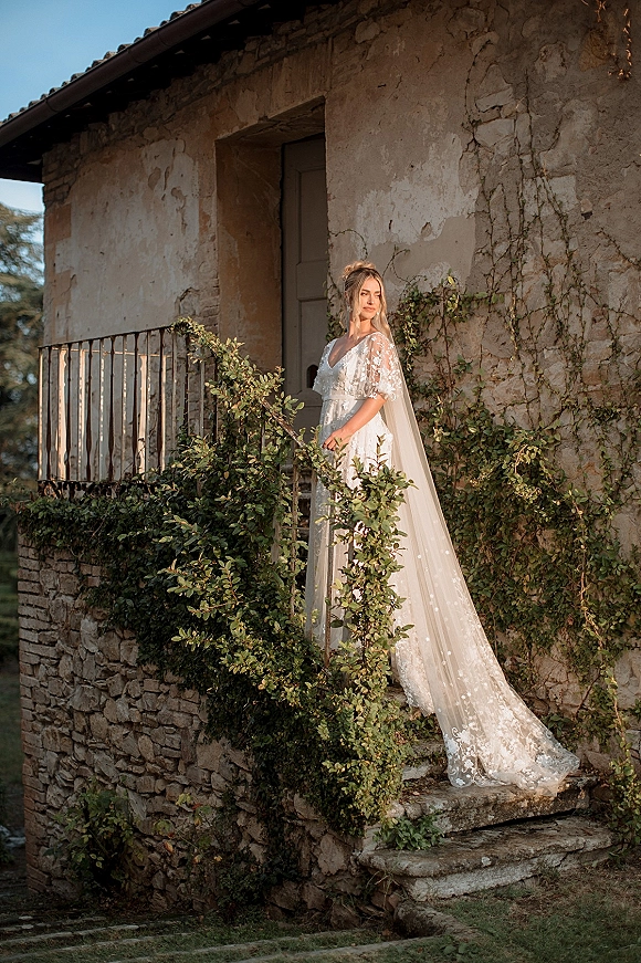 Bridal portrait of a bride in a lace wedding dress with a long veil, standing on stone steps by a vine-covered balcony under blue sky