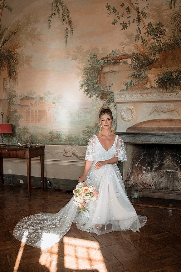 Bridal portrait of a bride sitting holding a rose and greenery bouquet in a lace deep V gown by a mural and marble fireplace, candle lanterns