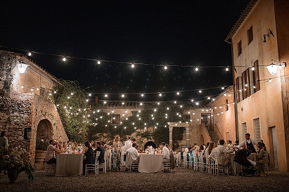 Outdoor wedding reception with string lights over long banquet tables, candle centerpieces, and white chairs in an ivy-walled courtyard at night