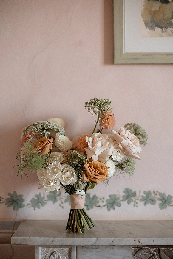 Bridal bouquet with neutral wedding bouquet tones of roses and dahlias, lace-wrapped stems and greenery on a marble mantel by a pink wall