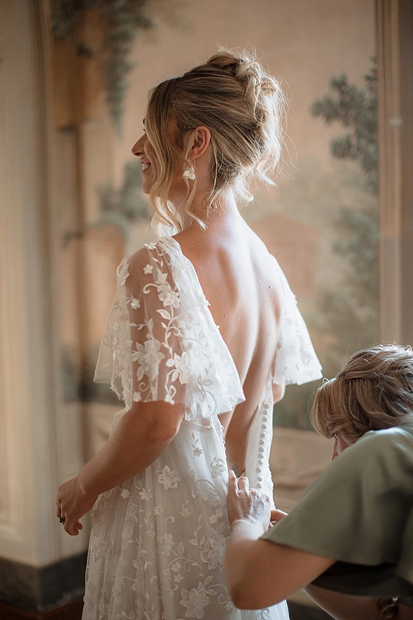 Bridal getting ready as bride gets dressed in a lace open-back gown with buttons, flutter sleeves and drop earrings in soft window light