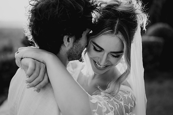 Wedding couple portrait of bride and groom embrace, groom whispering as she smiles in a bridal veil against a softly blurred outdoor landscape