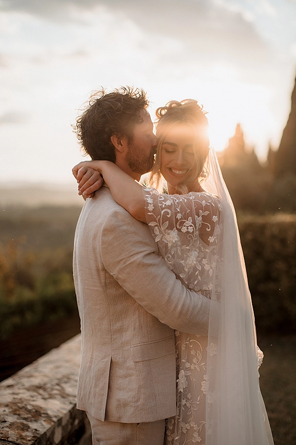 Couple portrait of bride and groom embrace as he kisses her forehead, veil and lace sleeves backlit by golden hour sun flare near a stone wall
