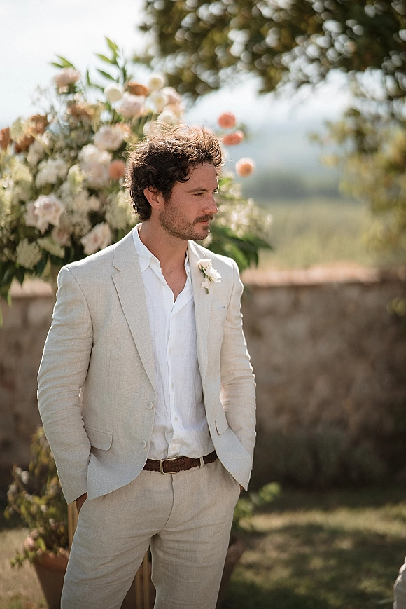 Groom portrait of a man in a light beige linen suit with boutonniere, hands in pockets before peach florals and a stone wall garden backdrop