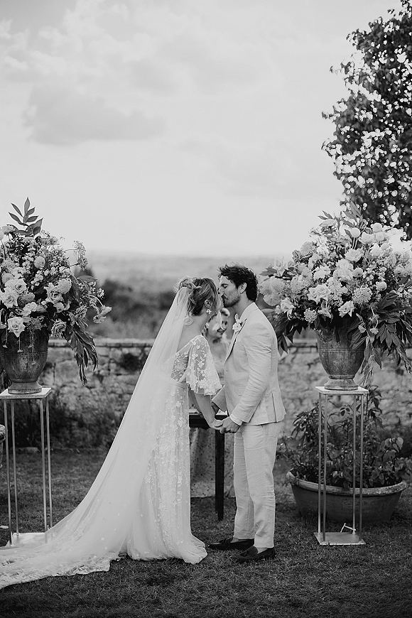Wedding kiss portrait of bride and groom kissing, bride in lace dress and veil, holding hands between floral pedestals on lawn by stone wall