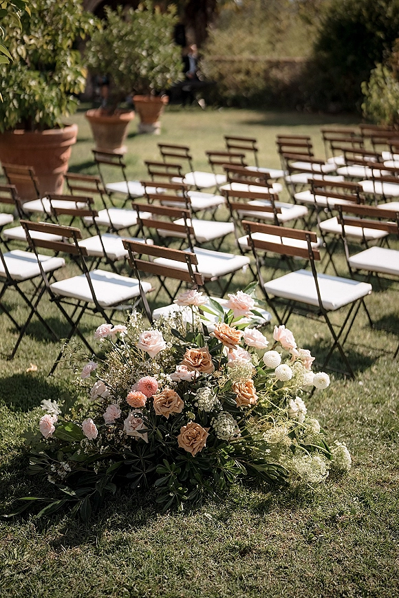Ceremony seating with wood folding chairs and white cushions in rows, featuring one meadow-style rose and greenery aisle arrangement on a lawn