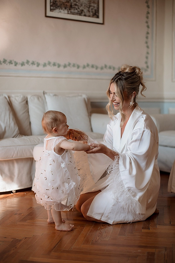 Bride with baby in a white satin robe with feather trim, holding a toddler in an embroidered tulle dress by a beige sofa