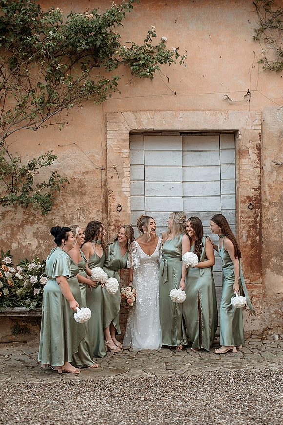 Bridesmaids portrait with bride in sage satin dresses holding bouquets, posed by a rustic stucco wall and vintage wooden door with vines