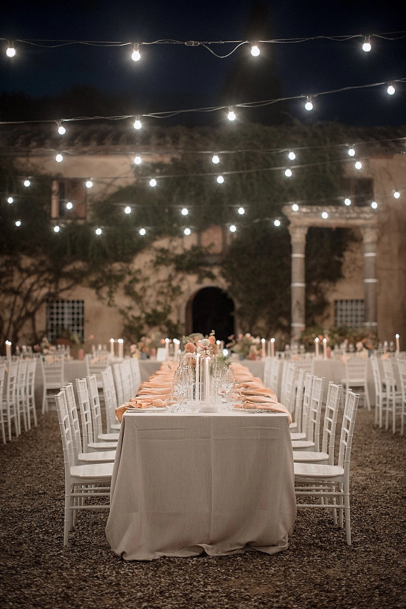Reception tablescape for an outdoor wedding reception on a long banquet table with taper candles under string lights in a courtyard