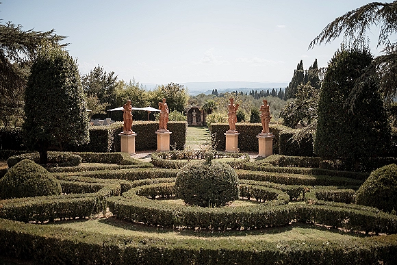 Garden wedding venue with classical statues and trimmed hedges surrounding a stone fountain, patio umbrellas on a manicured lawn with hills beyond
