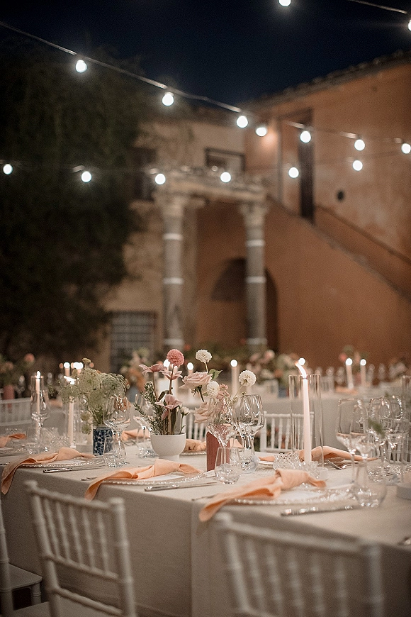 Reception tablescape with white linens, peach napkins, taper candles and bud-vase florals on round tables under string lights in a courtyard
