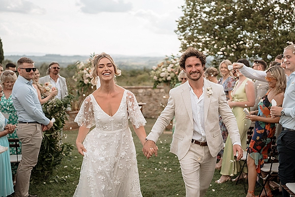 Wedding recessional as bride and groom walk the aisle holding hands while guests toss confetti petals in an outdoor garden setting
