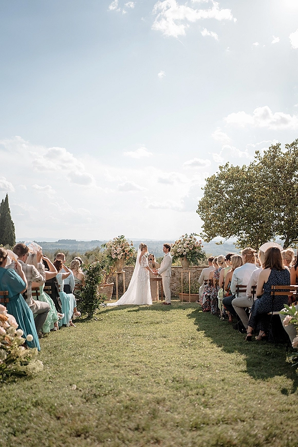 Wedding ceremony with bride and groom at a floral arch, bride in lace dress and veil on a green aisle set on a lawn by hills