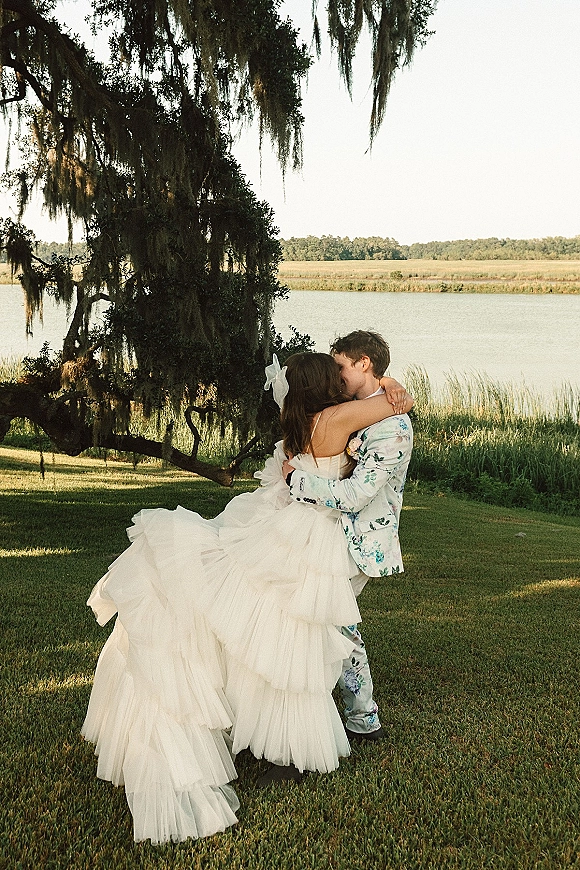 Wedding kiss portrait of bride and groom kissing as he dips her, strapless ruffle gown and floral suit under an oak with hanging moss by the lake