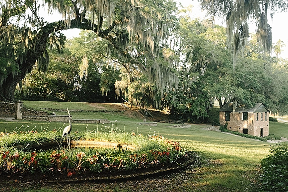 Wedding venue landscape with outdoor wedding venue grounds, oak trees draped in moss, flower beds, and a heron statue by a brick building in sunlight
