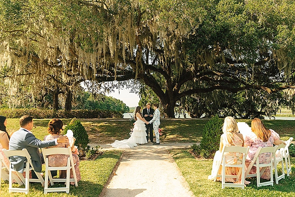 Wedding ceremony with bride in a ruffled gown and groom in patterned suit as officiant leads vows under oak trees with Spanish moss by a lake