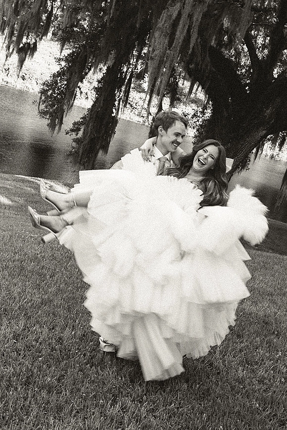 Couple portrait of groom carrying bride as she laughs, wedding dress ruffles flowing beside a river with mossy trees in back
