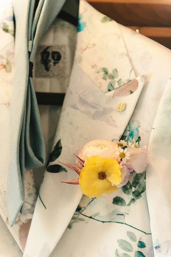Groom boutonniere with a yellow flower, white ranunculus and small daisies pinned to a floral suit lapel beside a light blue tie