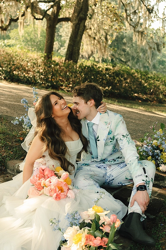 Couple portrait of bride and groom laughing in close embrace, her long veil and colorful bouquet against a garden path with Spanish moss