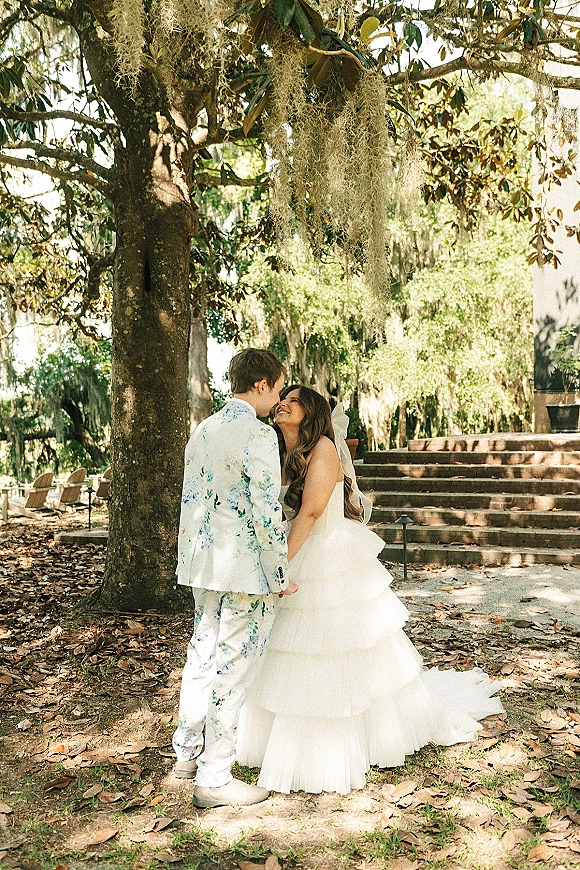 Couple portrait of bride and groom holding hands, bride looking up at groom under a mossy tree by garden stone steps