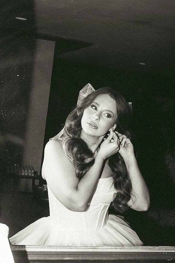 Bridal portrait of a bride putting on earrings in a strapless wedding dress, looking away in a dark interior with ceiling lights by a doorway