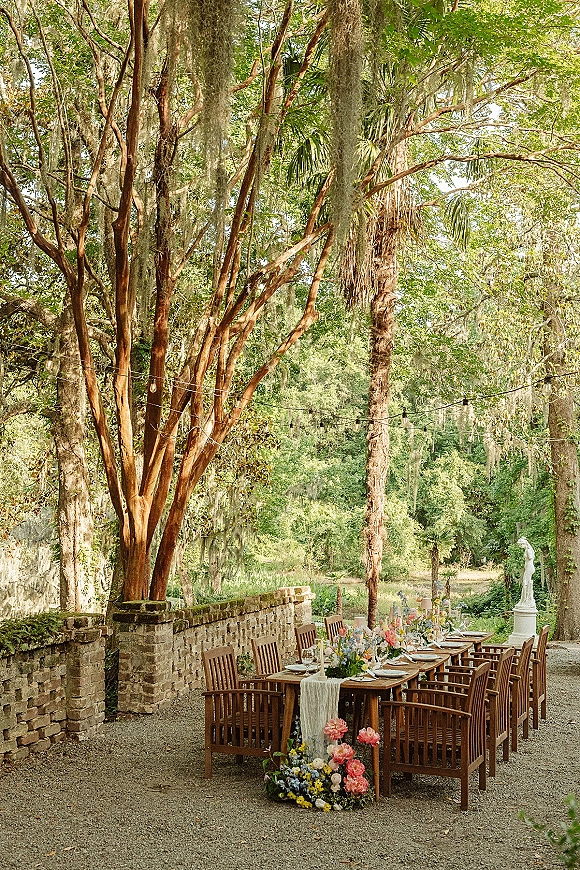 Outdoor reception tablescape with garden wedding reception details on a wood farm table, floral runner, and string lights under Spanish moss trees