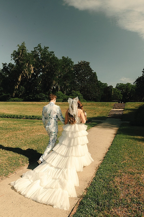 Couple portrait of newlyweds walking away down a garden path, bride’s long train and bow veil trailing behind under cloudy sky