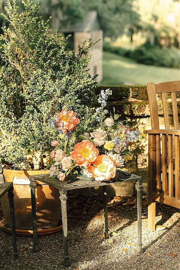 Wedding flowers coral peony centerpiece with blush roses and blue delphinium on a metal side table beside a wooden chair on a gravel patio