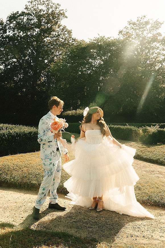 Couple portrait in a garden at golden hour, bride twirling her tulle skirt as groom in a floral suit holds a bouquet on a dirt path