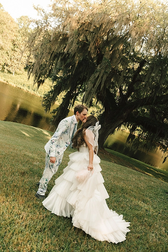 Wedding kiss by a lake, bride in a ruffled gown with bridal bow veil and groom in a floral suit beneath a mossy tree canopy